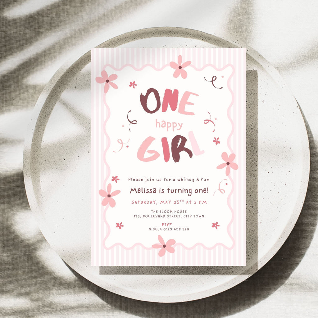 A pink and white 'One Happy Girl' birthday invitation card with floral illustrations, styled flat-lay on a round concrete plate with soft leaf shadows.