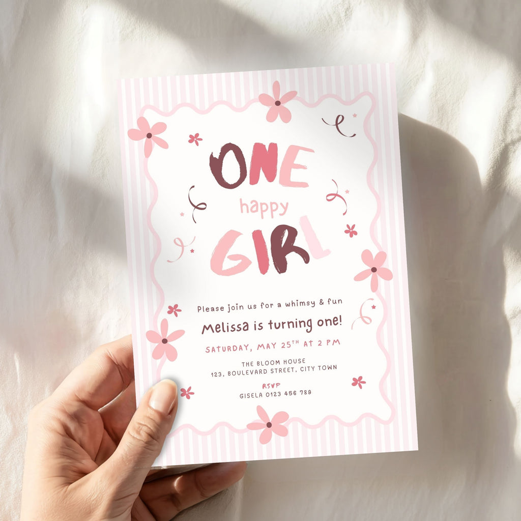 A hand holding a pink and white 'One Happy Girl' 1st birthday invitation card with floral designs, set against a soft, sunlit white fabric background.