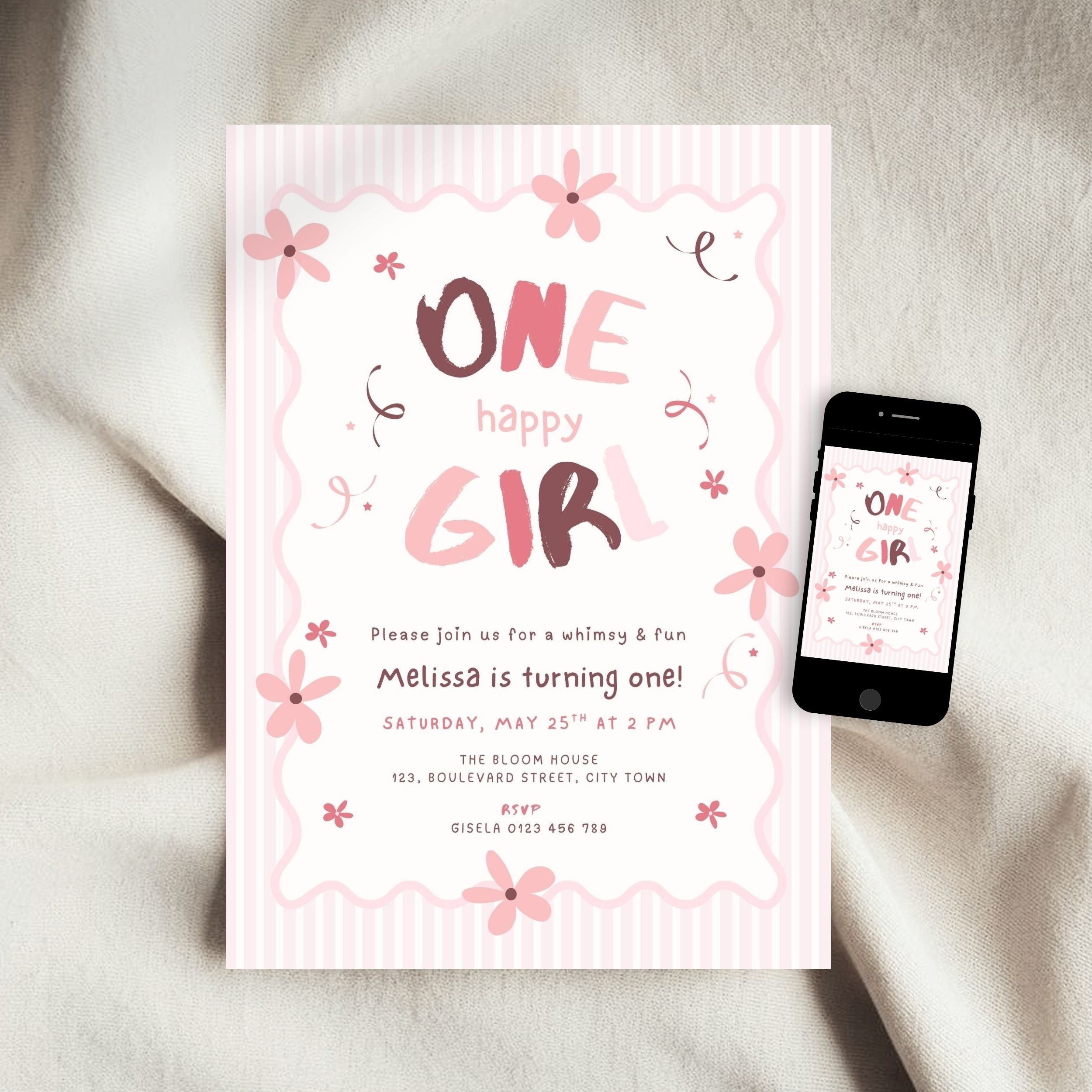 A pink and white striped 'One Happy Girl' first birthday invitation card is displayed on a soft beige fabric background next to a smartphone showing the digital version of the design.