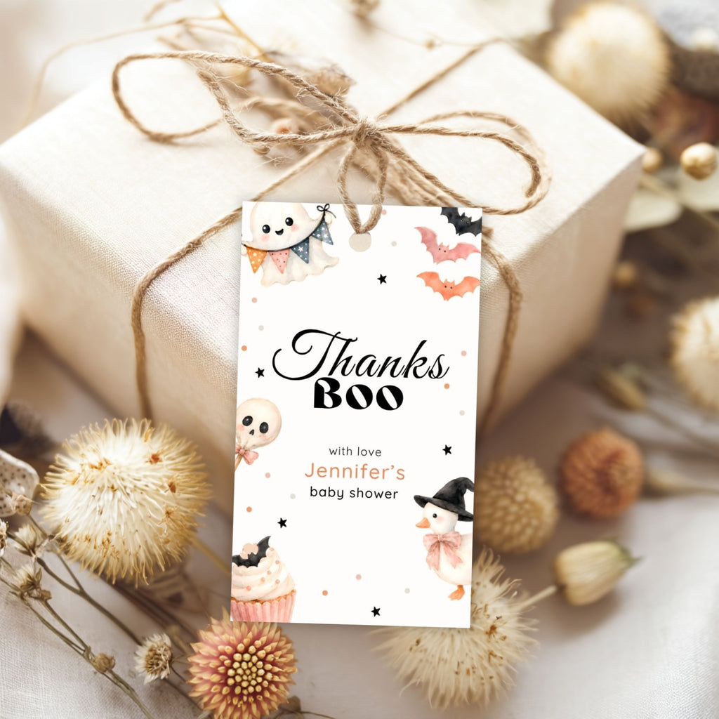 A personalized Halloween-themed baby shower gift tag reading 'Thanks Boo with love Jennifer's baby shower' tied to a wrapped gift box with twine, surrounded by dried flowers on a light fabric background.