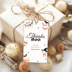 A personalized Halloween-themed baby shower gift tag reading 'Thanks Boo with love Jennifer's baby shower' tied to a wrapped gift box with twine, surrounded by dried flowers on a light fabric background.