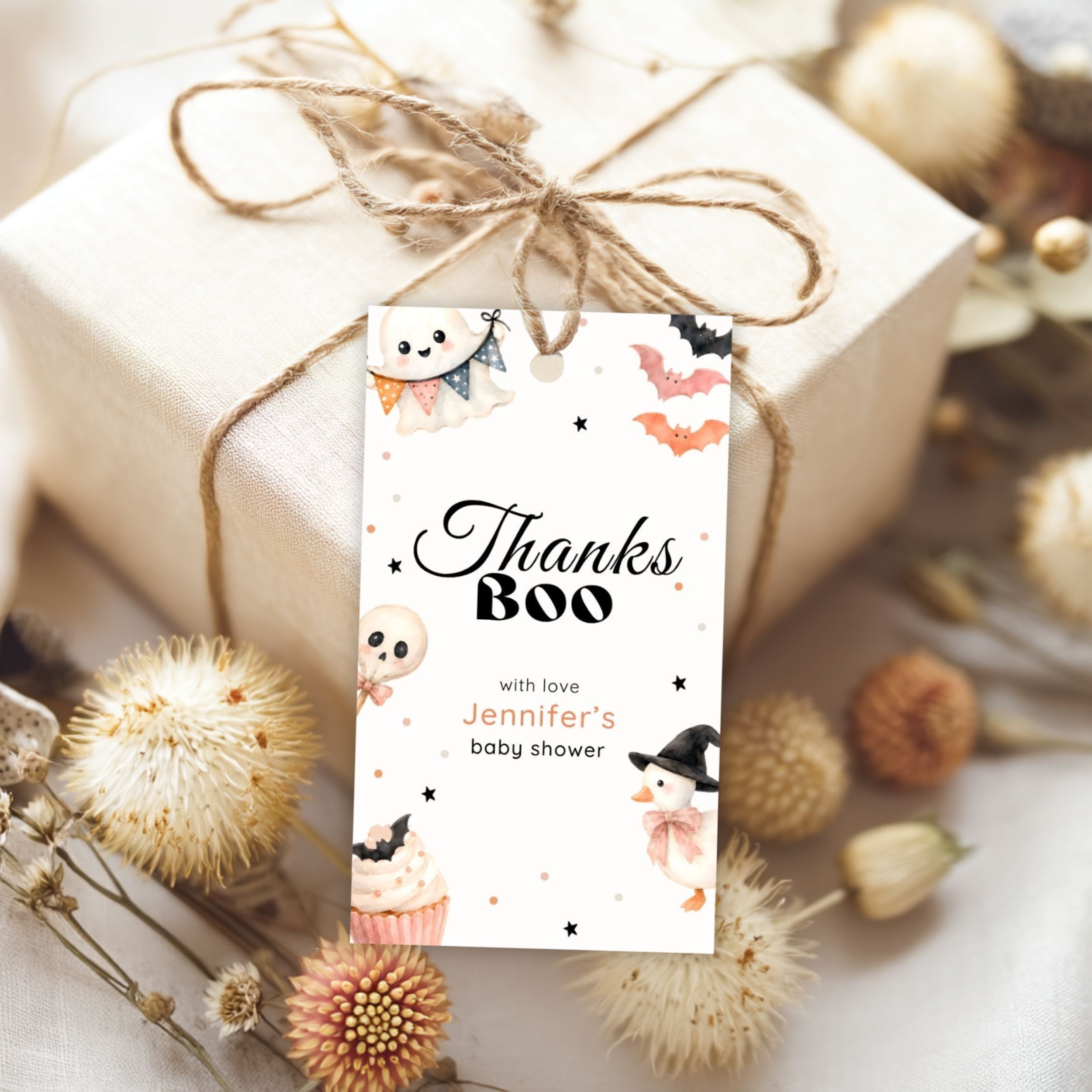 A personalized Halloween-themed baby shower gift tag reading 'Thanks Boo with love Jennifer's baby shower' tied to a wrapped gift box with twine, surrounded by dried flowers on a light fabric background.