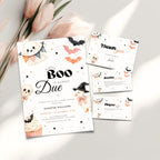 A Halloween baby shower invitation suite styled on a light neutral surface with pink flowers. The main product is a white invitation card featuring black text and cute illustrations of a ghost, cupcake, witch, bats, and stars, alongside a thank-you card, books for baby and a diaper raffle card.