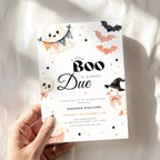 A hand holds a Halloween little boo is almost due baby shower invitation card with cute ghost, bat, pumpkin, and witch illustrations on a white background with soft shadows.