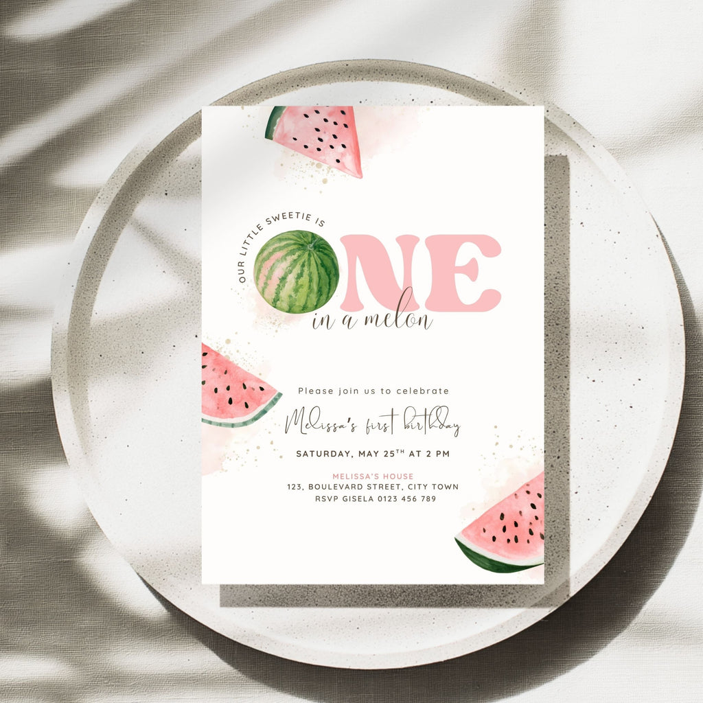 A watercolor-style first birthday invitation card featuring pink 'ONE in a millen' text and watermelon illustrations, styled on a speckled ceramic plate with soft shadows.