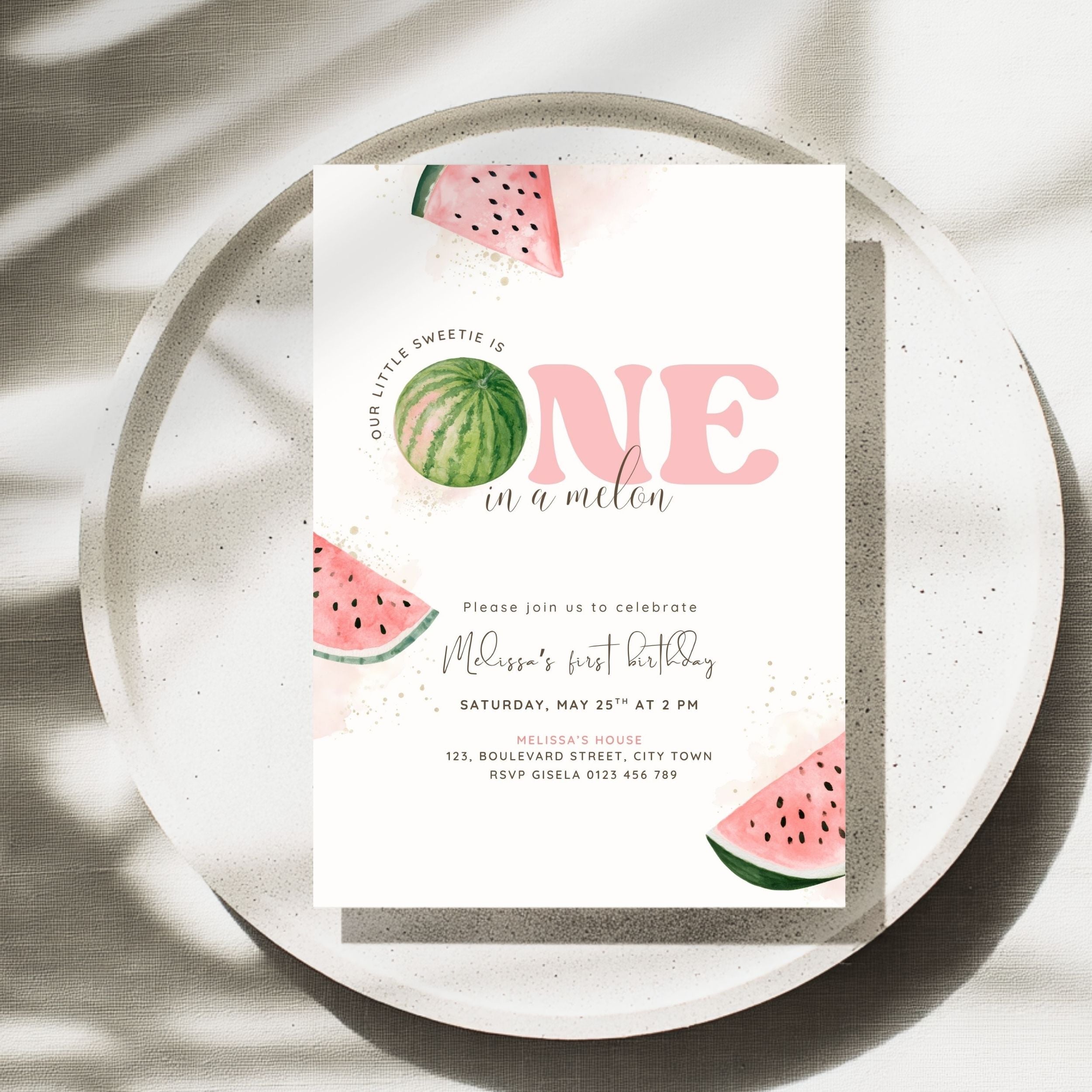 A watercolor-style first birthday invitation card featuring pink 'ONE in a millen' text and watermelon illustrations, styled on a speckled ceramic plate with soft shadows.