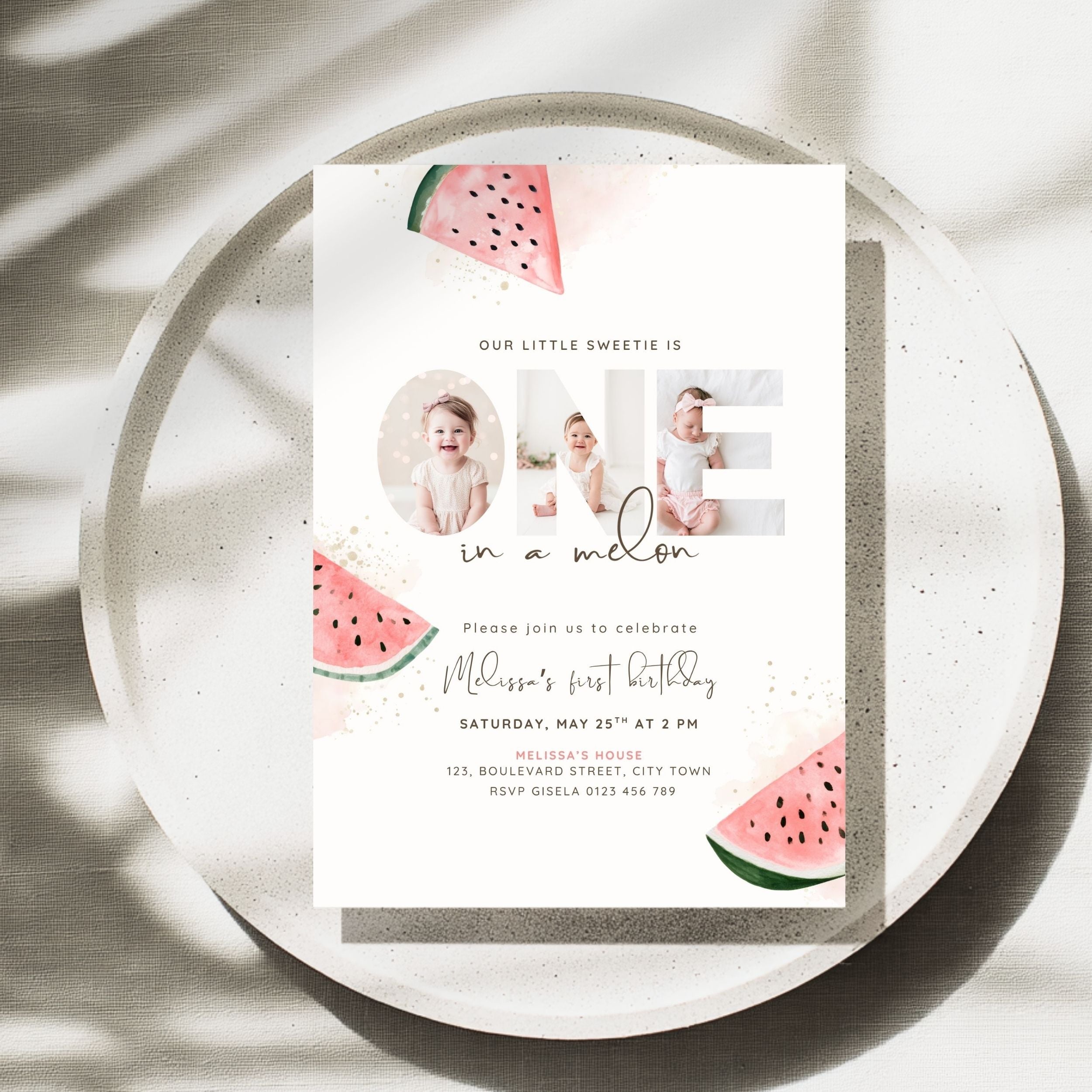 A watermelon-themed first birthday invitation card is displayed on a speckled ceramic plate with soft leaf shadows. The white card features illustrations of pink watermelon slices and photos of a baby in three circular frames, with the word 'ONE' stylized to include the baby photos.