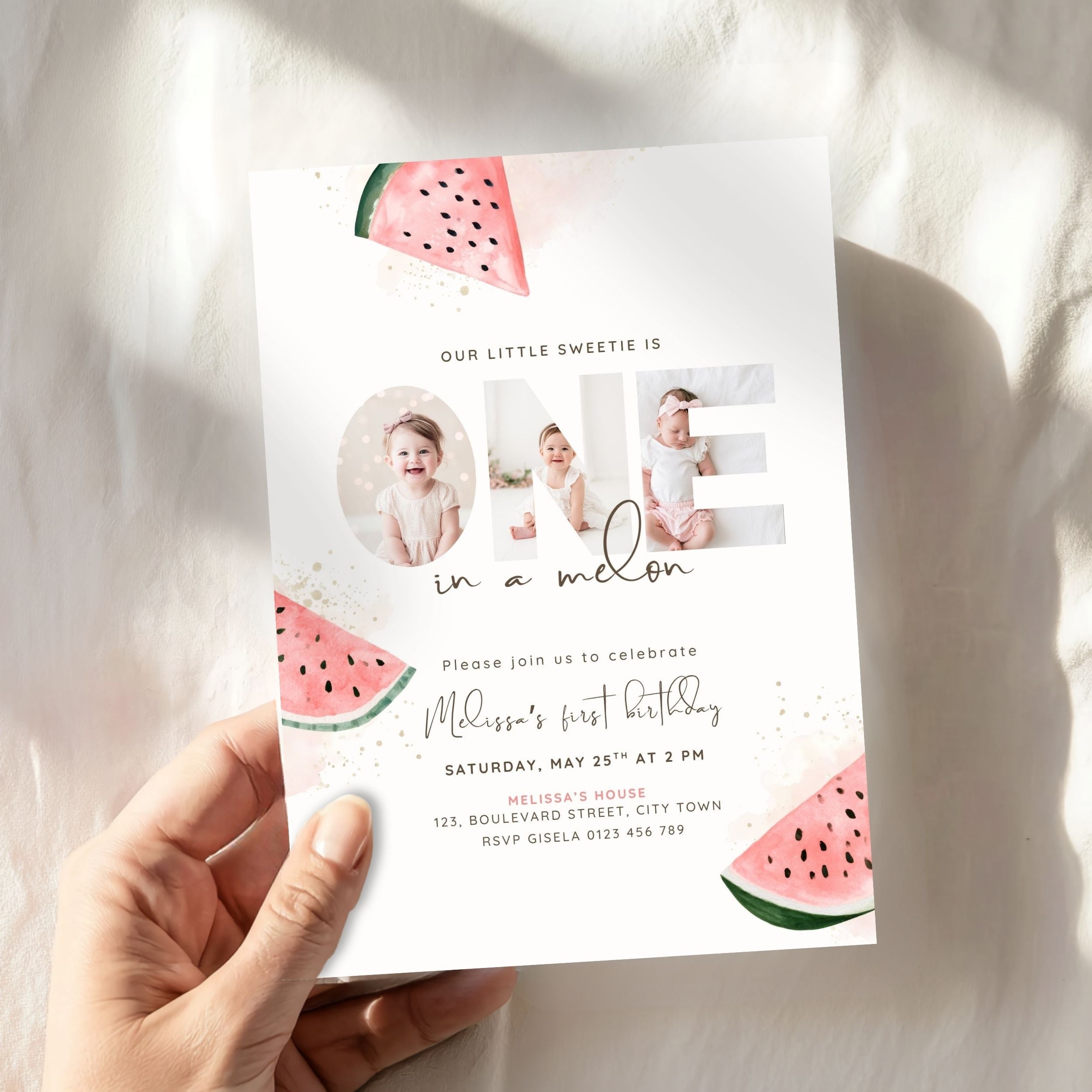 A hand holding a custom watermelon-themed first birthday invitation card against a softly lit, textured white fabric background.