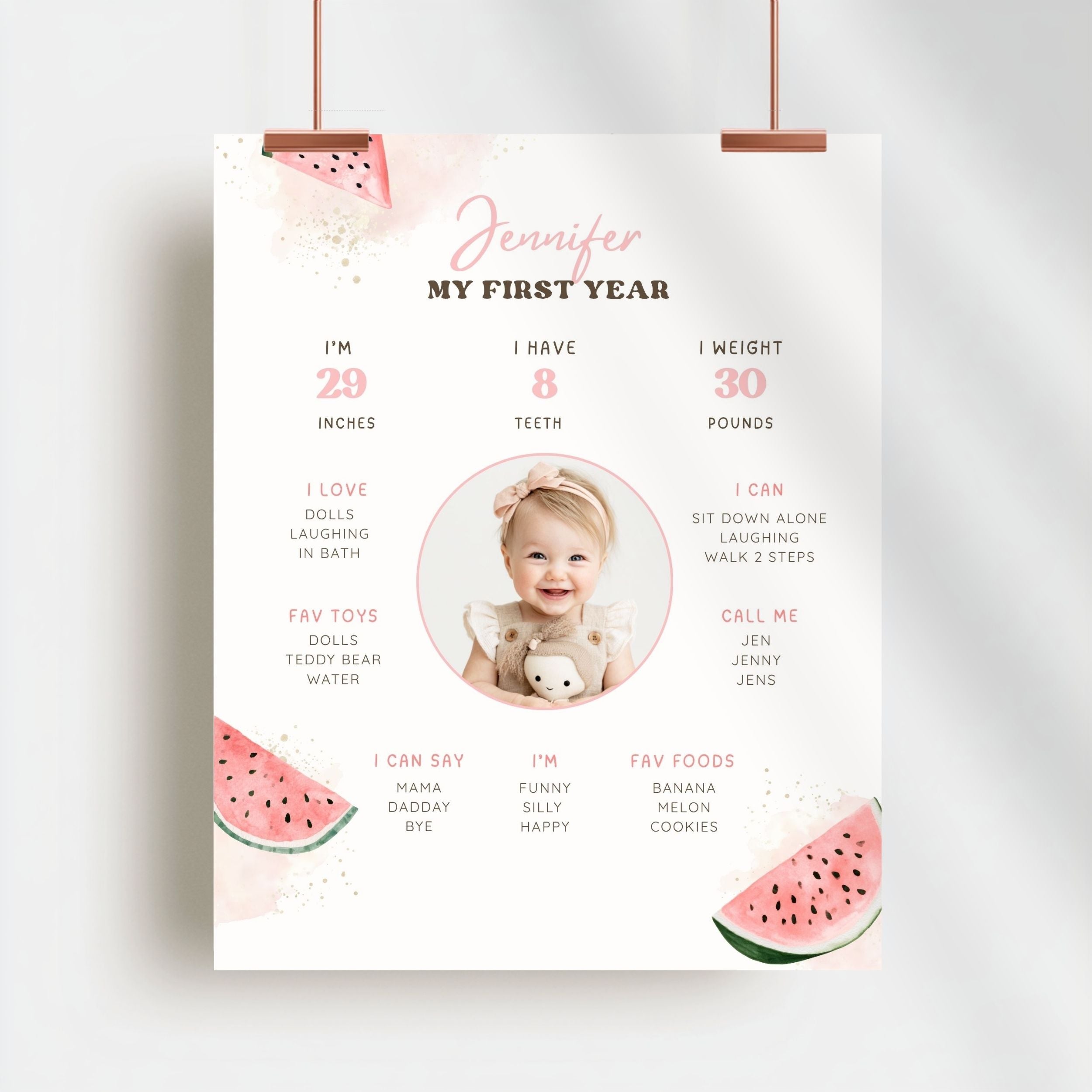 A personalized first birthday milestone poster for a child named Jennifer, featuring watermelon illustrations and a circular photo placeholder of a baby. The poster is shown hanging from two copper-colored clips against a light gray background.