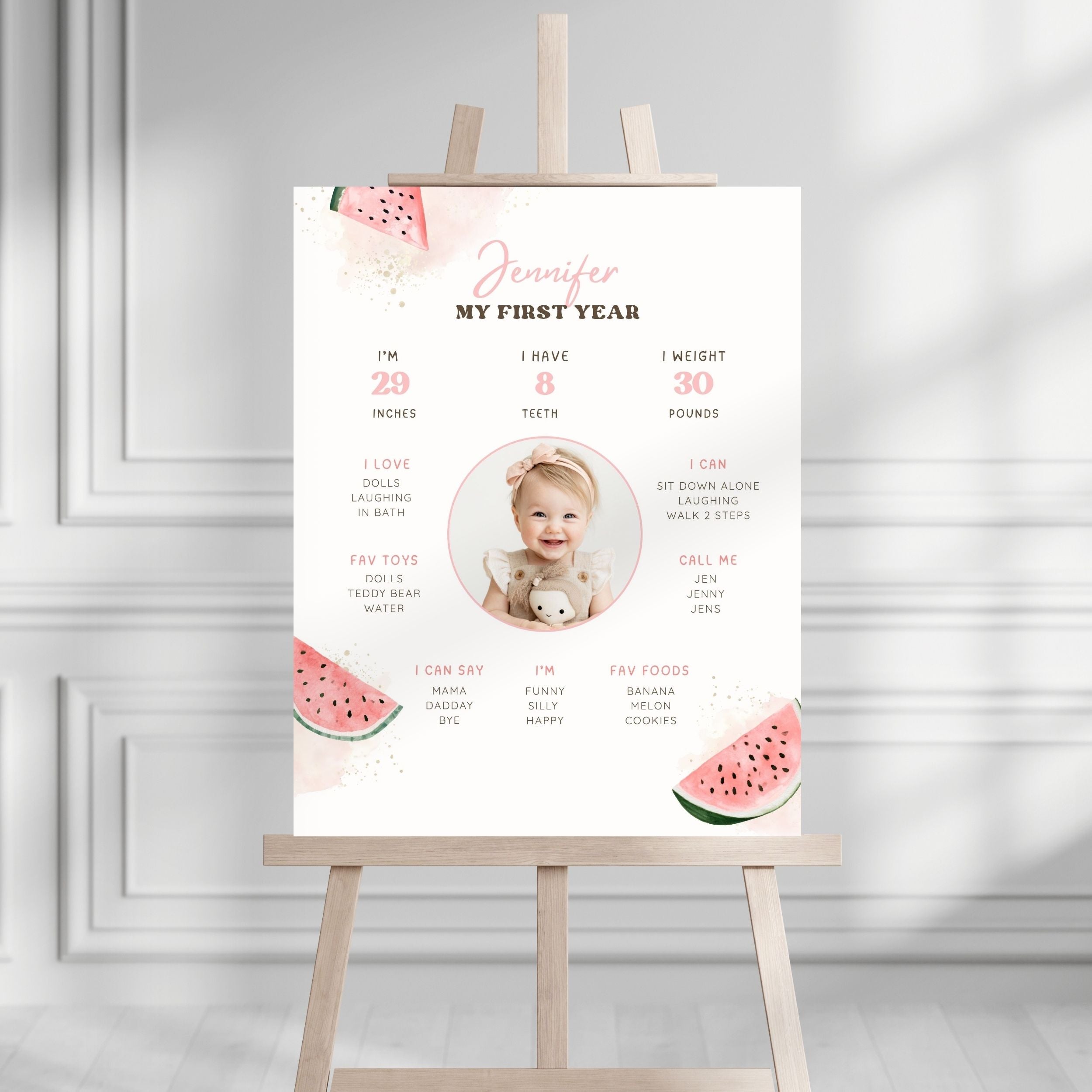 A personalized baby milestone poster for 'Jennifer' displayed on a wooden easel. The white poster features pink and green watermelon graphics, a central photo placeholder of a baby, and cute text detailing baby milestones in a clean, modern room setting.