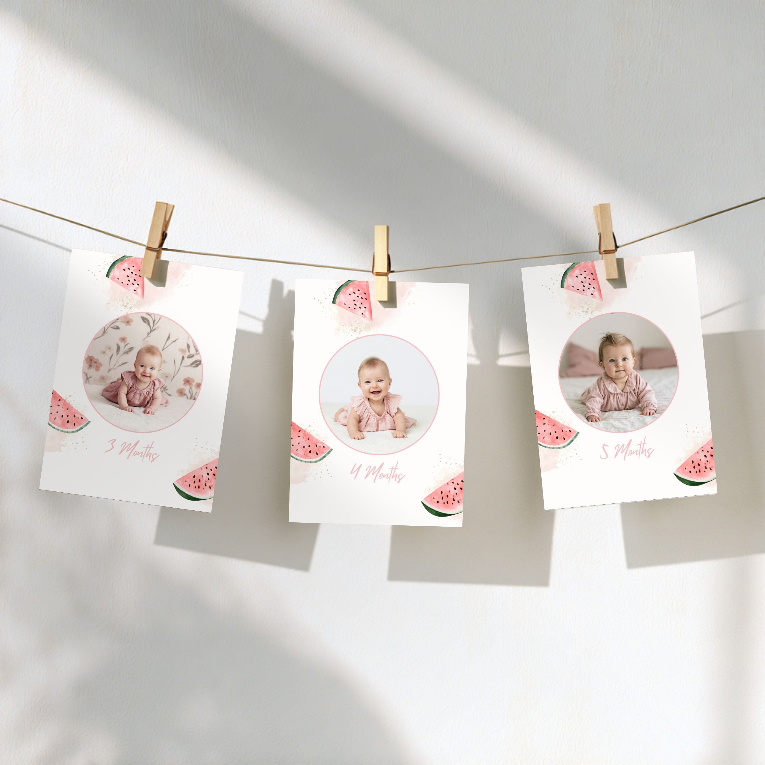 Three white baby milestone photos hanging from a string with wooden clothespins against a white wall. Each card has a circular photo of a baby, watermelon slice illustrations, and the date '5 Months' written in pink script.