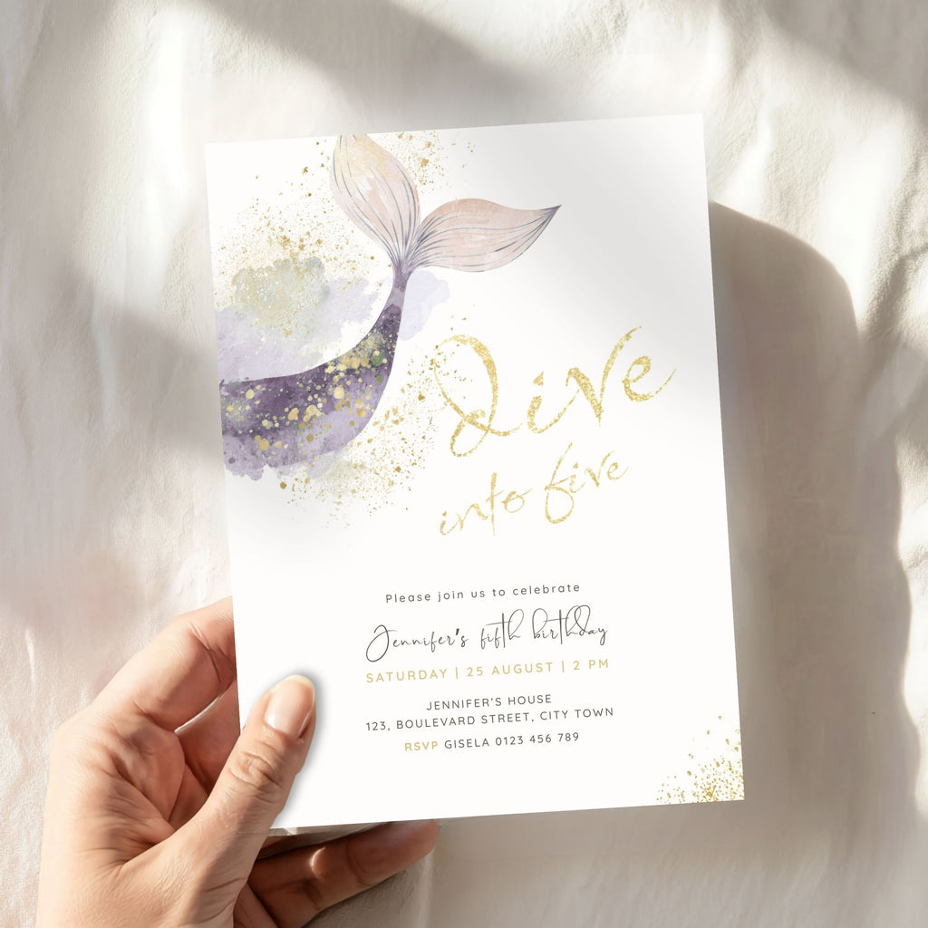 A hand holds a white 'Dive into Five' mermaid-themed birthday invitation card featuring a purple and gold watercolor mermaid tail, gold glitter accents, and gold foil-style lettering, set against a softly lit white fabric background.