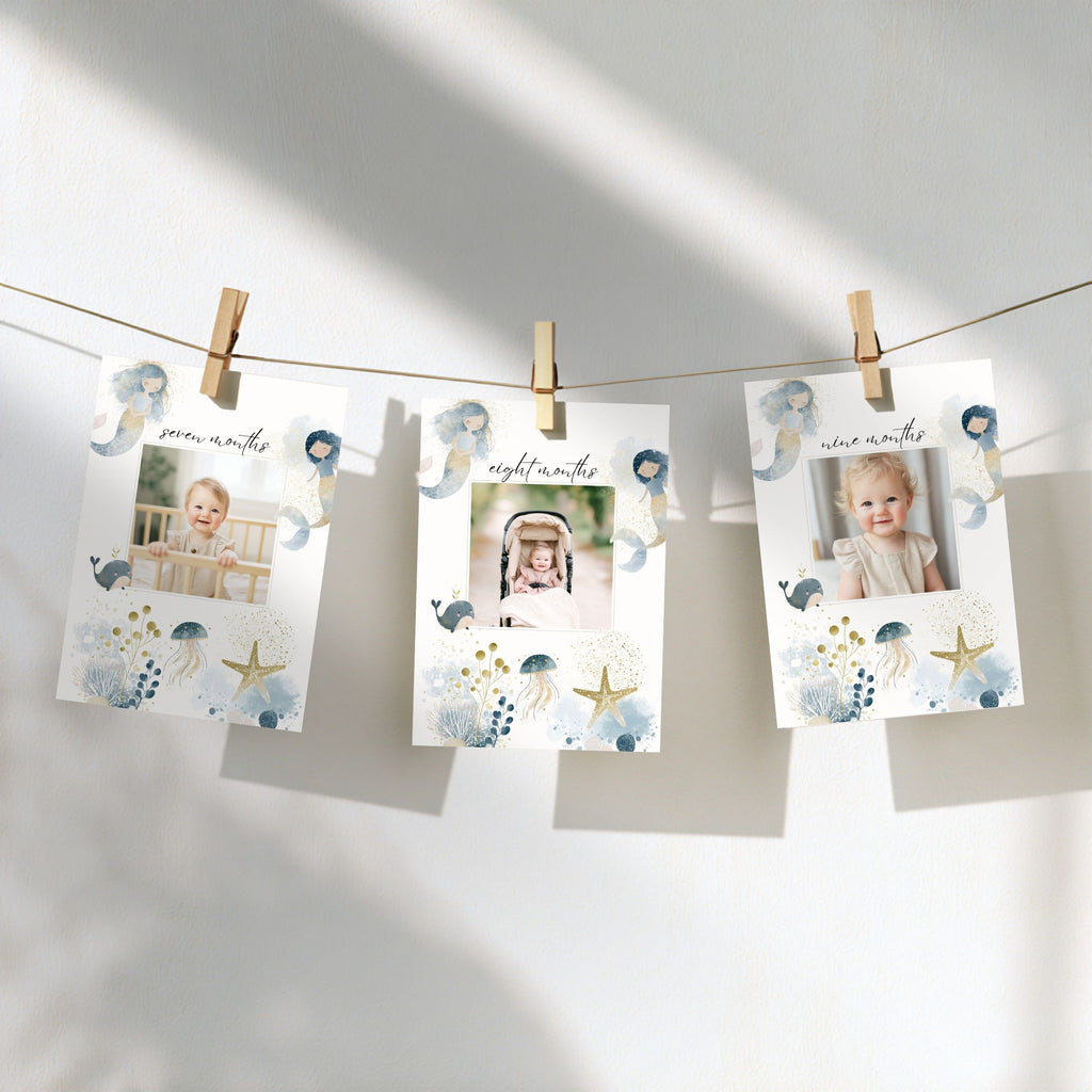 Three printed milestone photos hang from a string with wooden clothespins against a white wall. The cards have a soft blue and gold seashell and starfish illustration design, with baby photos shown in the center slots.
