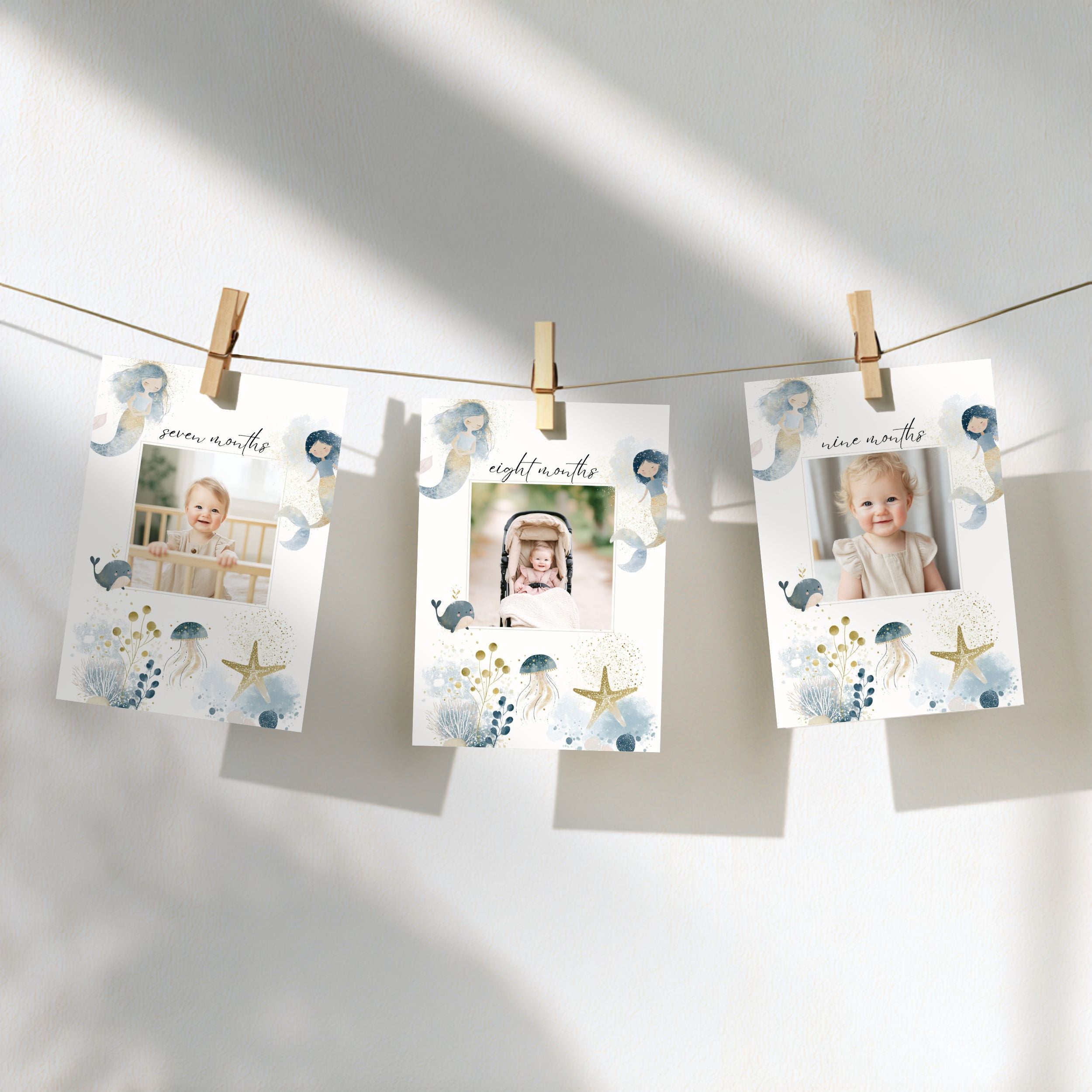 Three printed milestone photos hang from a string with wooden clothespins against a white wall. The cards have a soft blue and gold seashell and starfish illustration design, with baby photos shown in the center slots.