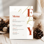 A vertical christmas menu card displayed upright on a light fabric surface, surrounded by decorative pinecones and dried grass in a soft neutral setting. The card has a warm off-white background with large rust-red serif letters 'E' and 'Y' and small gold abstract line accents, plus a printed menu title 'Menu' and small course text.