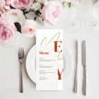 A christmas menu card with elegant typography and gold monogram details, styled on a white plate with silver cutlery and pink roses in the background.