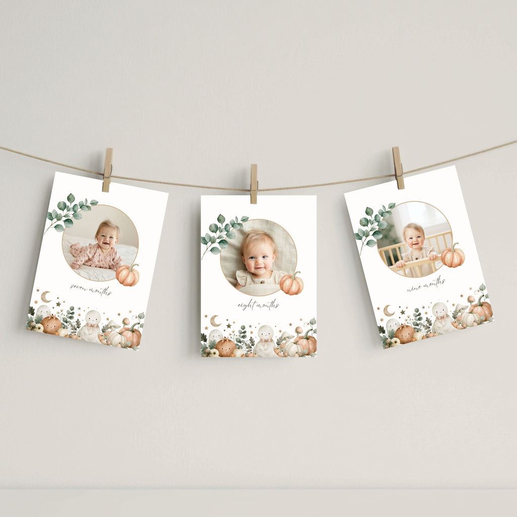 Three vertical baby milestone photo cards with autumn watercolor designs are clipped to a string against a neutral wall. Each card features a circular photo of a baby, a pumpkin, eucalyptus leaves, and cute animal illustrations at the bottom.