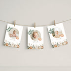 Three vertical baby milestone photo cards with autumn watercolor designs are clipped to a string against a neutral wall. Each card features a circular photo of a baby, a pumpkin, eucalyptus leaves, and cute animal illustrations at the bottom.