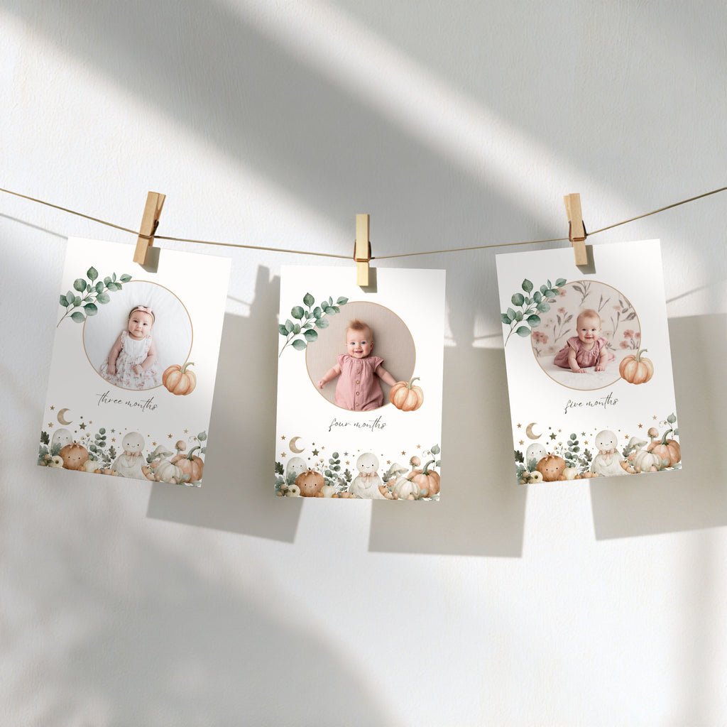 Three personalized baby milestone cards hang from a string with wooden clothespins against a white wall. Each card features a circular photo of a baby, a small pumpkin illustration, and a watercolor autumn border with leaves and pumpkins.