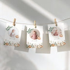 Three personalized baby milestone cards hang from a string with wooden clothespins against a white wall. Each card features a circular photo of a baby, a small pumpkin illustration, and a watercolor autumn border with leaves and pumpkins.