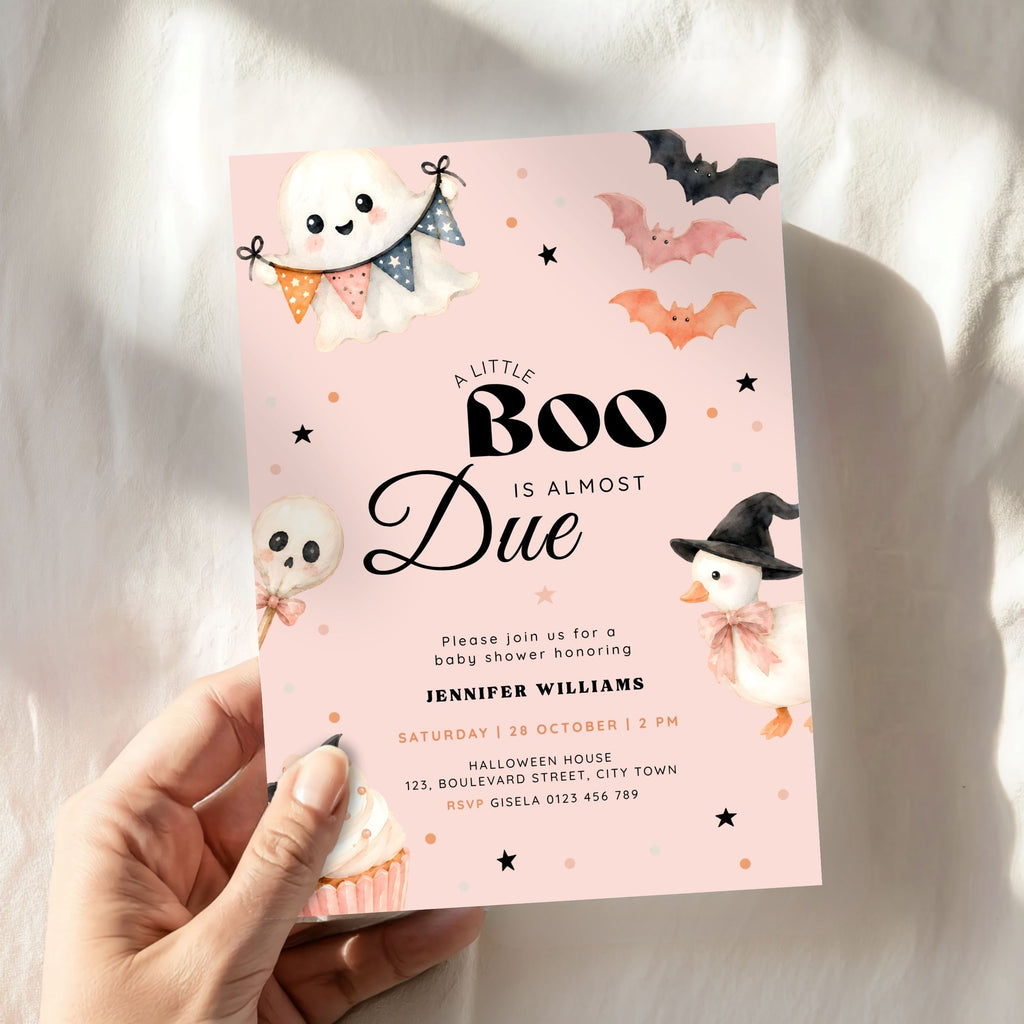A hand holds a pink Halloween baby shower invitation card with a light blue background, featuring cute illustrations of a ghost, witch duck, bats, and cupcakes. The card is photographed against a soft white fabric backdrop with warm sunlight and shadows.