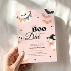 A hand holds a pink Halloween baby shower invitation card with a light blue background, featuring cute illustrations of a ghost, witch duck, bats, and cupcakes. The card is photographed against a soft white fabric backdrop with warm sunlight and shadows.