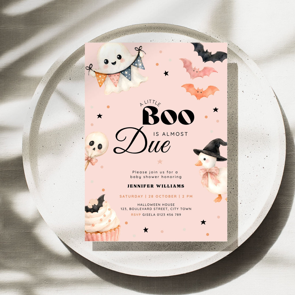 A pink Halloween-themed a little boo is alsmost due baby shower invitation card displayed on a round tray. The card has a light blue background with cute illustrations of a ghost, bats, an orange pumpkin, a witch duck, a cupcake, and black decorative stars.