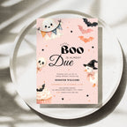 A pink Halloween-themed a little boo is alsmost due baby shower invitation card displayed on a round tray. The card has a light blue background with cute illustrations of a ghost, bats, an orange pumpkin, a witch duck, a cupcake, and black decorative stars.