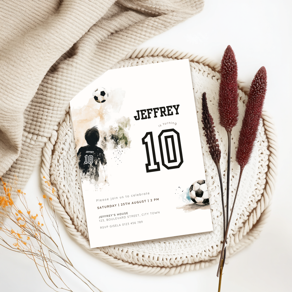 A custom soccer-themed 10th birthday invitation card styled in a flat lay with a beige knit blanket, a crocheted placemat, dried bunny tail grass, and small yellow wildflowers. The white card features black text reading 'JEFFREY 10' with watercolor soccer graphics.