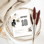 A custom soccer-themed 10th birthday invitation card styled in a flat lay with a beige knit blanket, a crocheted placemat, dried bunny tail grass, and small yellow wildflowers. The white card features black text reading 'JEFFREY 10' with watercolor soccer graphics.