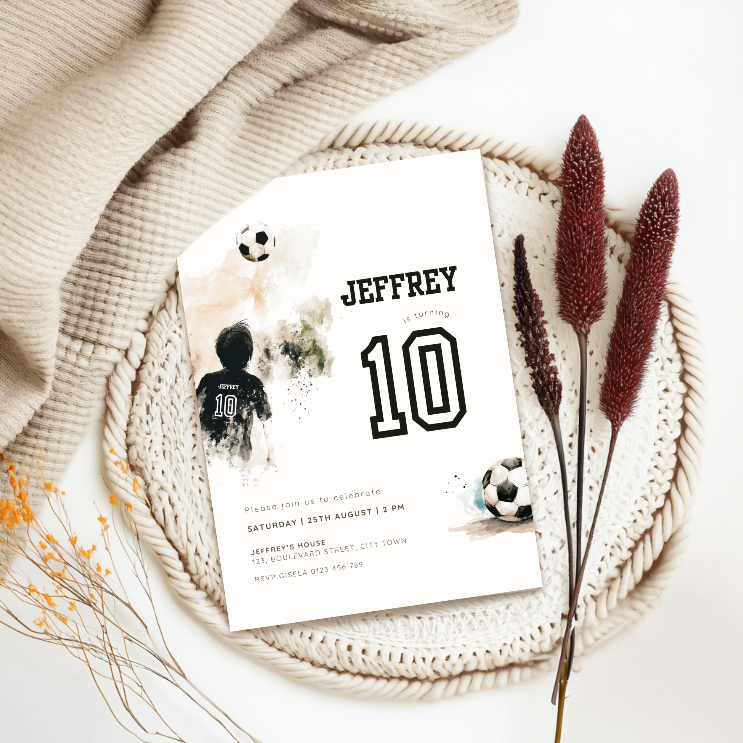 A custom soccer-themed 10th birthday invitation card styled in a flat lay with a beige knit blanket, a crocheted placemat, dried bunny tail grass, and small yellow wildflowers. The white card features black text reading 'JEFFREY 10' with watercolor soccer graphics.