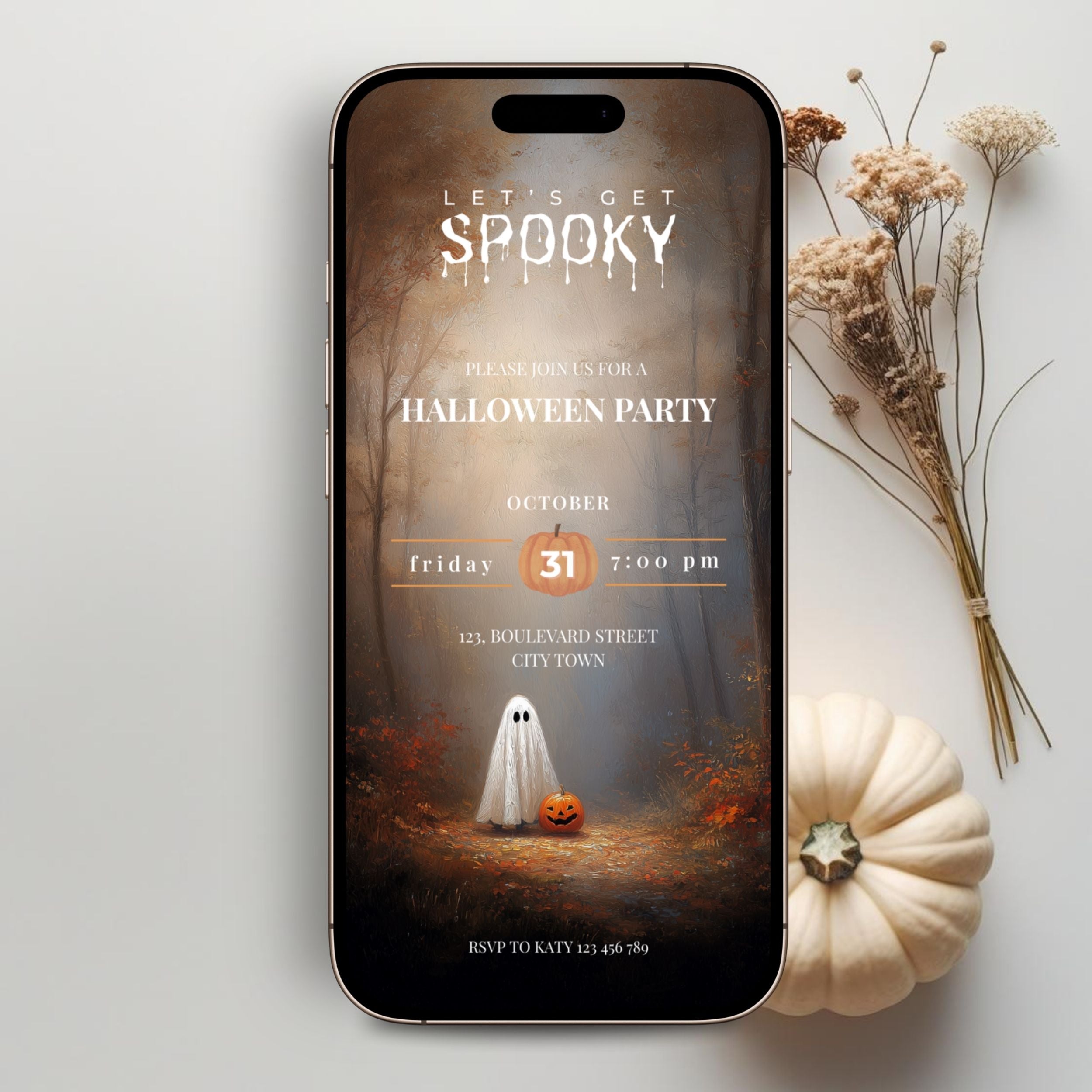 A smartphone centered on a light gray surface, displaying a Halloween party invitation design with a ghost, pumpkin, and autumn forest scene. Dried flowers and a small cream pumpkin are arranged around the phone.
