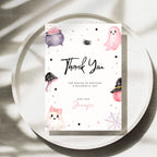 A Halloween-themed first birthday invitation card displayed on a speckled white plate. The card features cute illustrations of a pink ghost, purple cauldron, black spider, mushroom, witch hat, and spiderwebs on a white background with black and pink text.