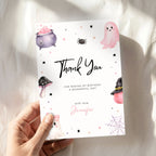 A hand holds a white Halloween-themed thank you card with cute illustrations of a purple cauldron, pink ghost, spider, mushroom, witch hat, and spiderweb. The card reads 'Thank You' and includes personalized text for 'Jennifer' against a soft white fabric background.