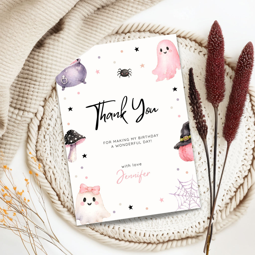 A Halloween-themed thank you card styled in a flat lay on a woven placemat with dried flowers and a cream knit blanket. The card features cute watercolor illustrations of ghosts, a spider, a mushroom, a witch hat, and a spiderweb in pink, purple, black, and white, with the text 'Thank You FOR MAKING MY BIRTHDAY A WONDERFUL DAY! with love Jennifer'.