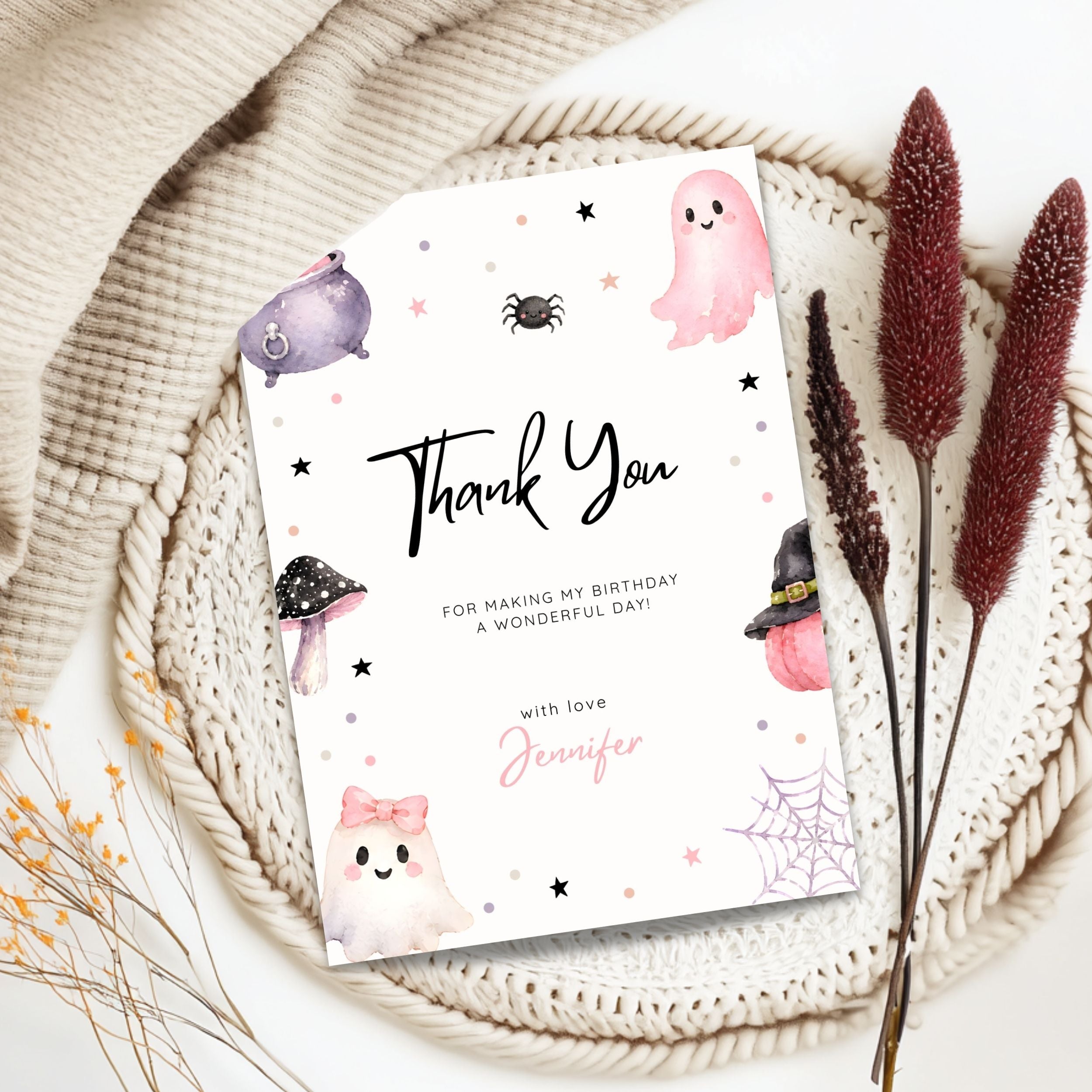 A Halloween-themed thank you card styled in a flat lay on a woven placemat with dried flowers and a cream knit blanket. The card features cute watercolor illustrations of ghosts, a spider, a mushroom, a witch hat, and a spiderweb in pink, purple, black, and white, with the text 'Thank You FOR MAKING MY BIRTHDAY A WONDERFUL DAY! with love Jennifer'.