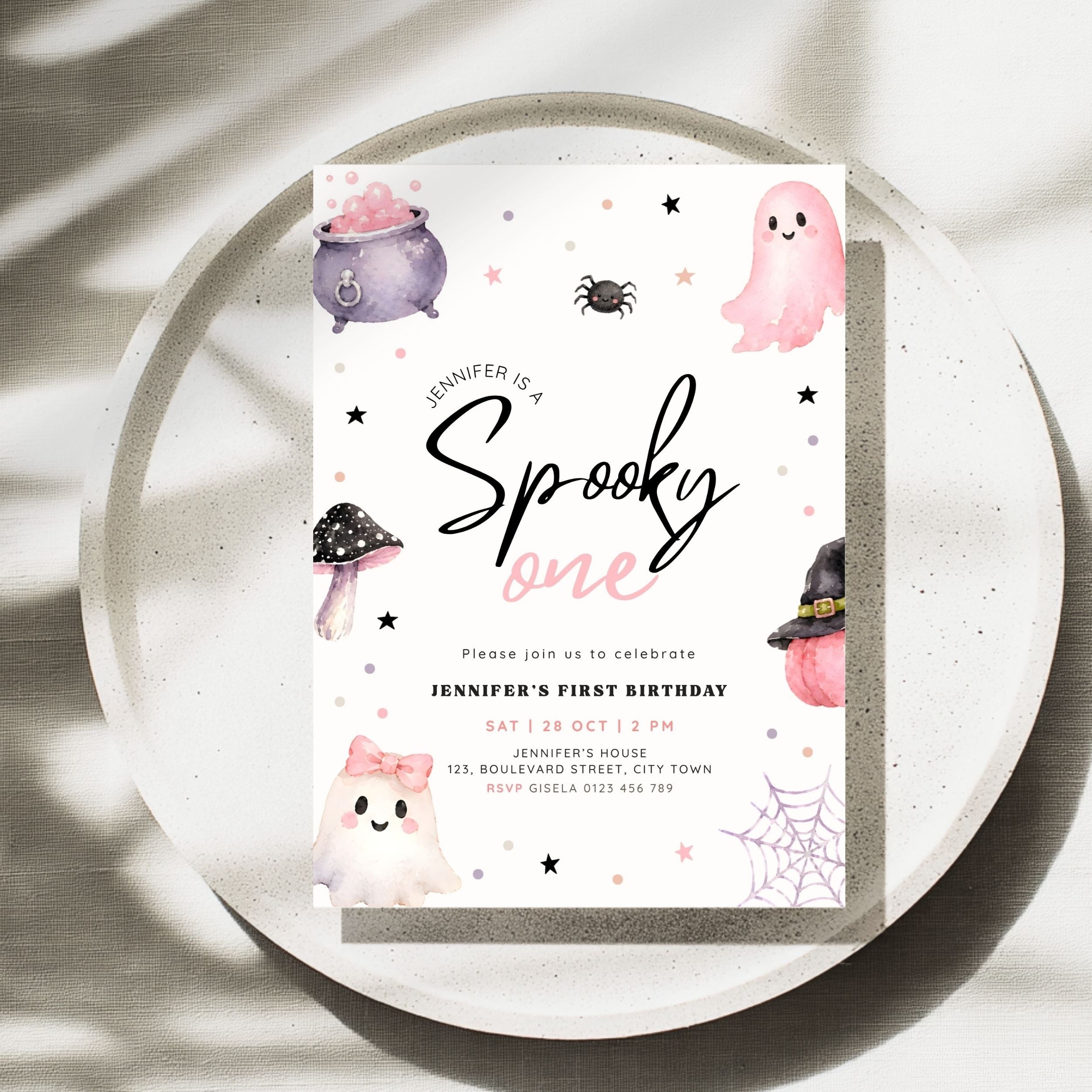 A Halloween-themed first birthday invitation card displayed on a speckled white plate. The card features cute illustrations of a pink ghost, purple cauldron, black spider, mushroom, witch hat, and spiderwebs on a white background with black and pink text.