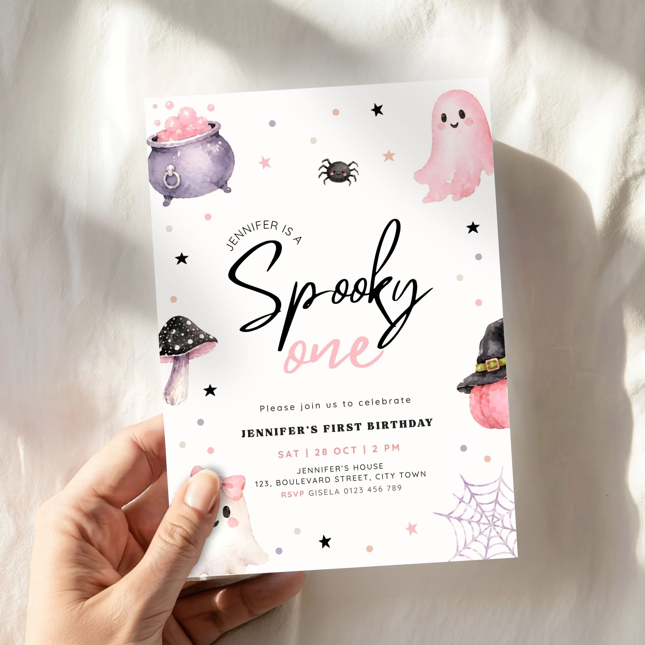 A hand holds a Halloween-themed first birthday invitation card against a soft white fabric background. The card features cute watercolor illustrations of a purple cauldron, pink ghost, black spider, purple mushroom, witch hat, spiderweb, and the text 'Jennifer is a Spooky one'.