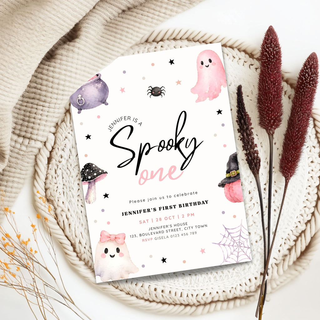 A Halloween-themed first birthday invitation card styled in a flat lay on a beige knit blanket with dried decorative grass stems. The card features cute watercolor illustrations of a pink ghost, a purple monster, a spider, mushrooms, and a witch hat on a white background.