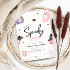 A Halloween-themed first birthday invitation card styled in a flat lay on a beige knit blanket with dried decorative grass stems. The card features cute watercolor illustrations of a pink ghost, a purple monster, a spider, mushrooms, and a witch hat on a white background.
