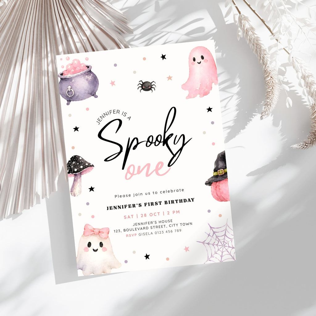 A Halloween-themed first birthday invitation card styled on a light gray surface with dried palm leaves. The card features cute watercolor illustrations of ghosts, a cauldron, a spider, a mushroom, and a witch hat on a white background with black and pink text.