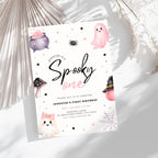 A Halloween-themed first birthday invitation card styled on a light gray surface with dried palm leaves. The card features cute watercolor illustrations of ghosts, a cauldron, a spider, a mushroom, and a witch hat on a white background with black and pink text.