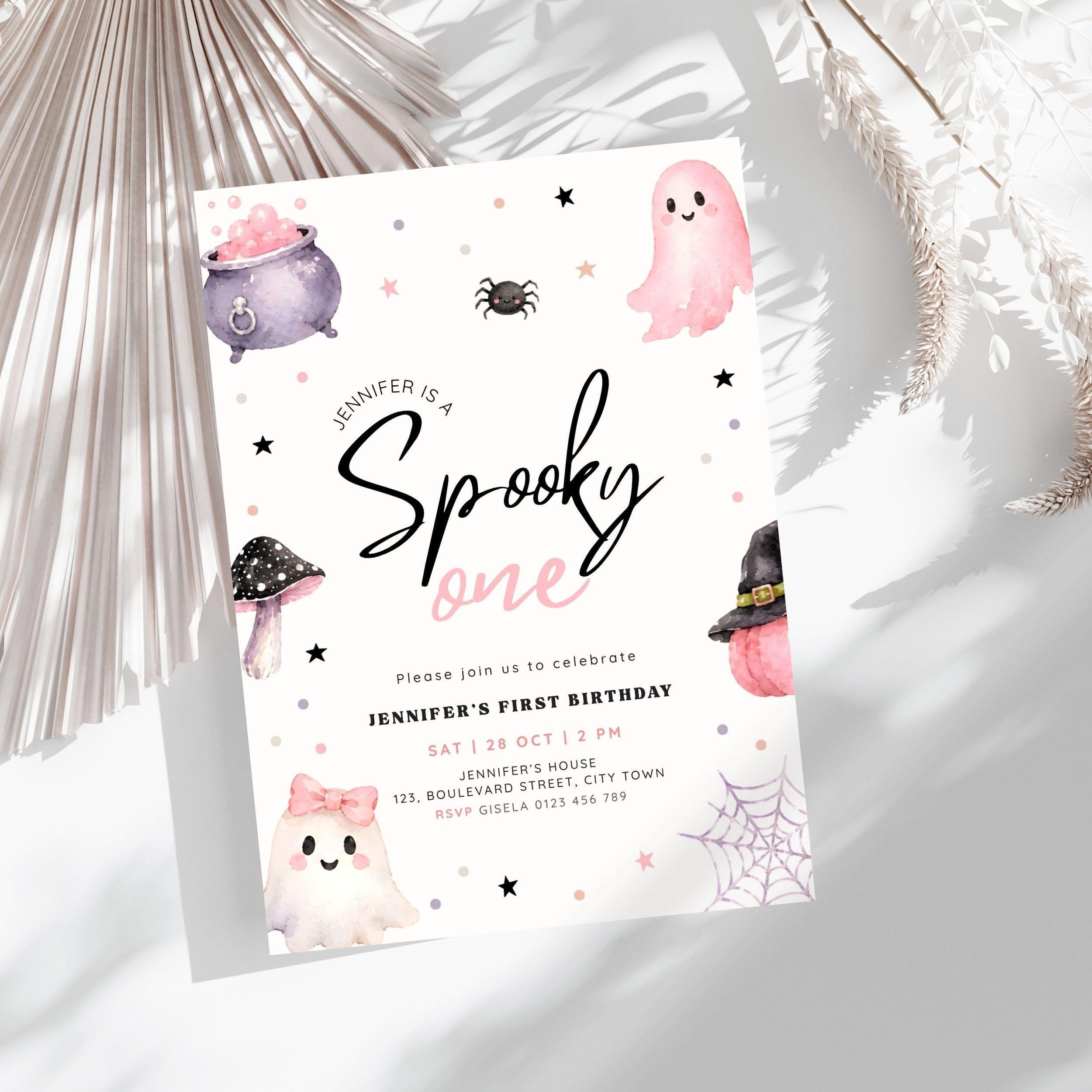 A Halloween-themed first birthday invitation card styled on a light gray surface with dried palm leaves. The card features cute watercolor illustrations of ghosts, a cauldron, a spider, a mushroom, and a witch hat on a white background with black and pink text.