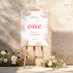 Sweet One Birthday welcome sign on an easel with floral decorations against a stone wall.