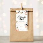 A kraft paper party favor bag with a wooden clip holding a white 'Thanks Boo' tag, shown against a softly blurred warm bokeh background.