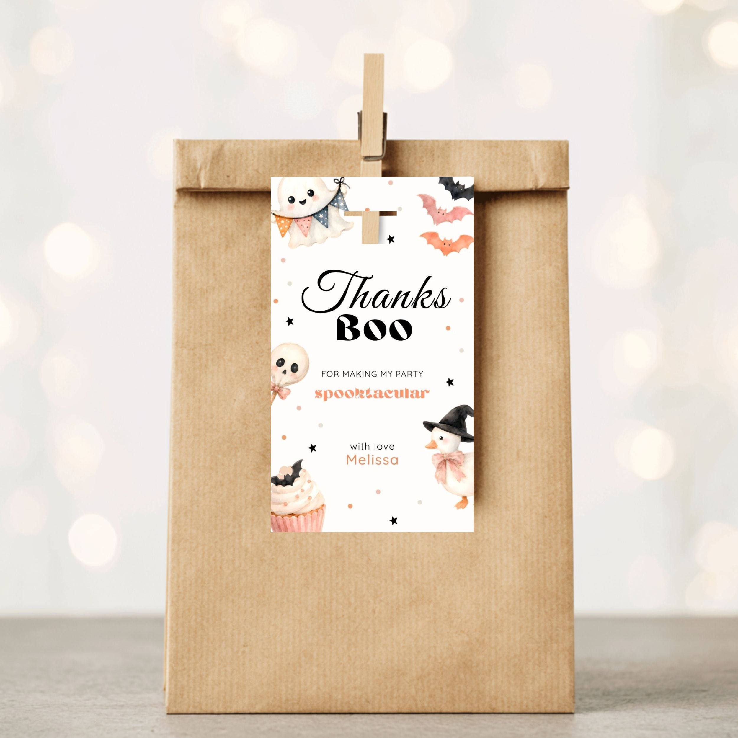 A kraft paper party favor bag with a wooden clip holding a white 'Thanks Boo' tag, shown against a softly blurred warm bokeh background.