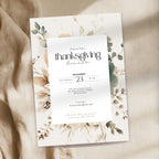 A Thanksgiving dinner invitation card with a white center panel and botanical floral border in muted beige, green, and brown tones, photographed on draped beige fabric.
