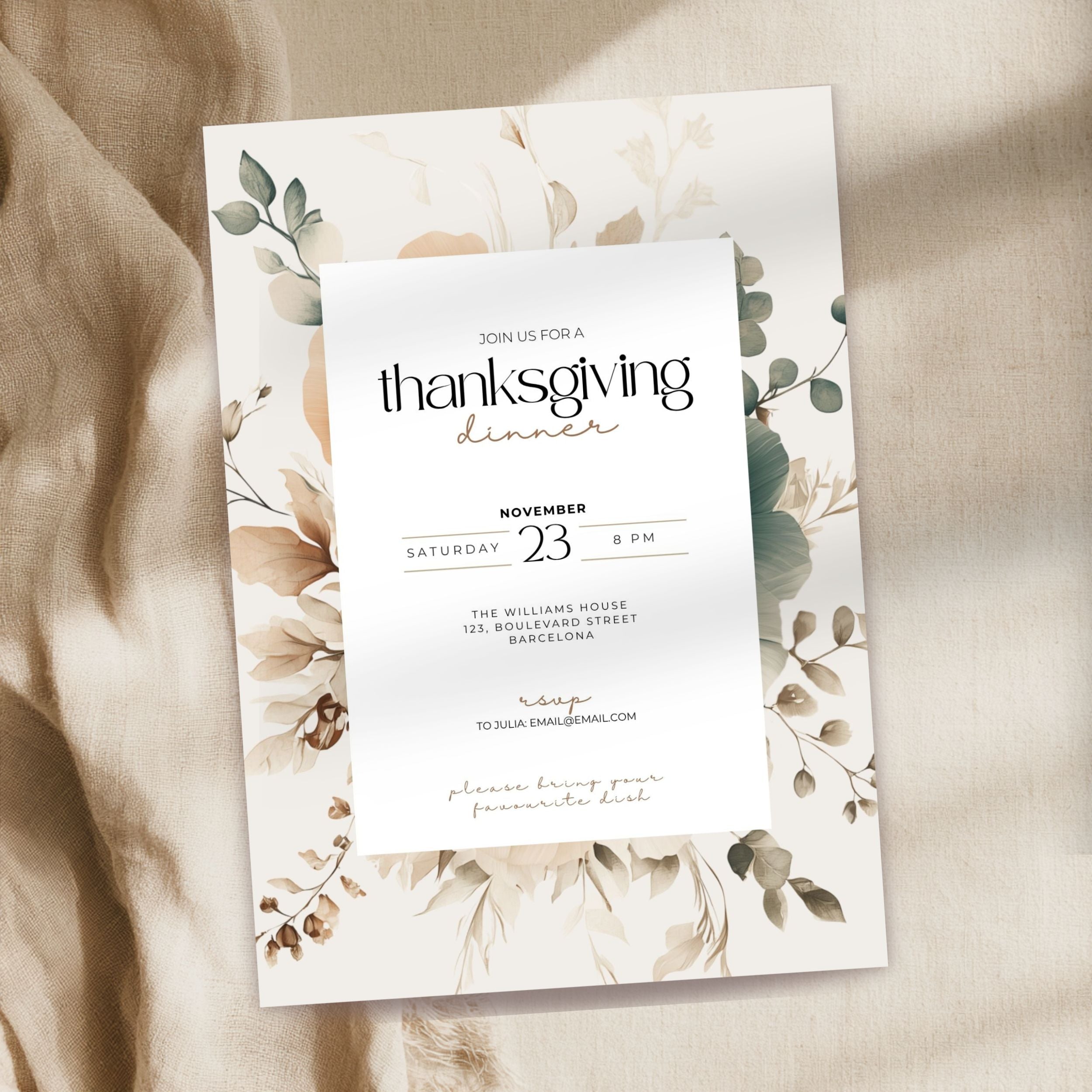 A Thanksgiving dinner invitation card with a white center panel and botanical floral border in muted beige, green, and brown tones, photographed on draped beige fabric.