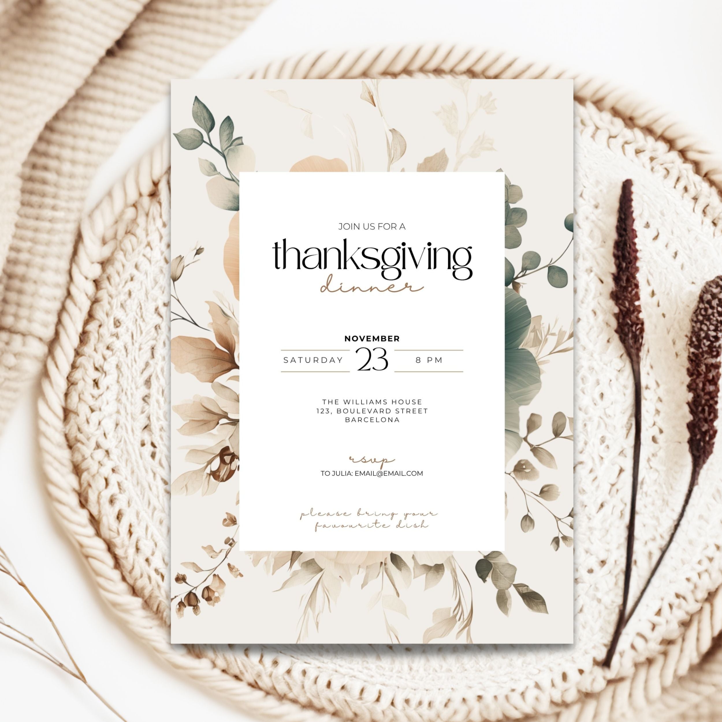 A Thanksgiving dinner invitation card with a white center panel and muted botanical border in beige, green, and brown tones, styled on a woven tray with dried stems.