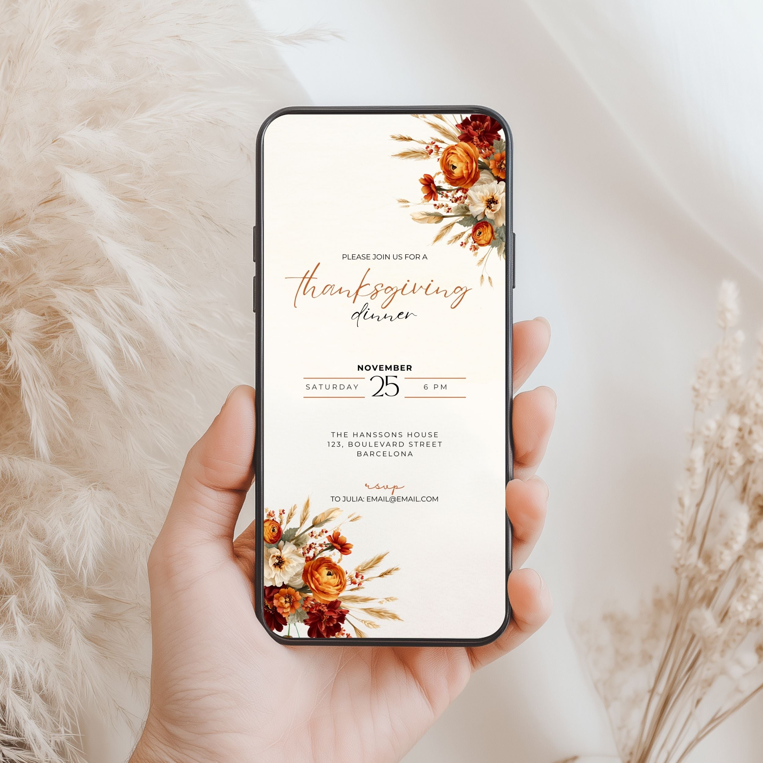 A hand holds a smartphone displaying a Thanksgiving dinner invitation design with floral illustrations in warm autumn tones, set against a soft beige background with dried grasses.